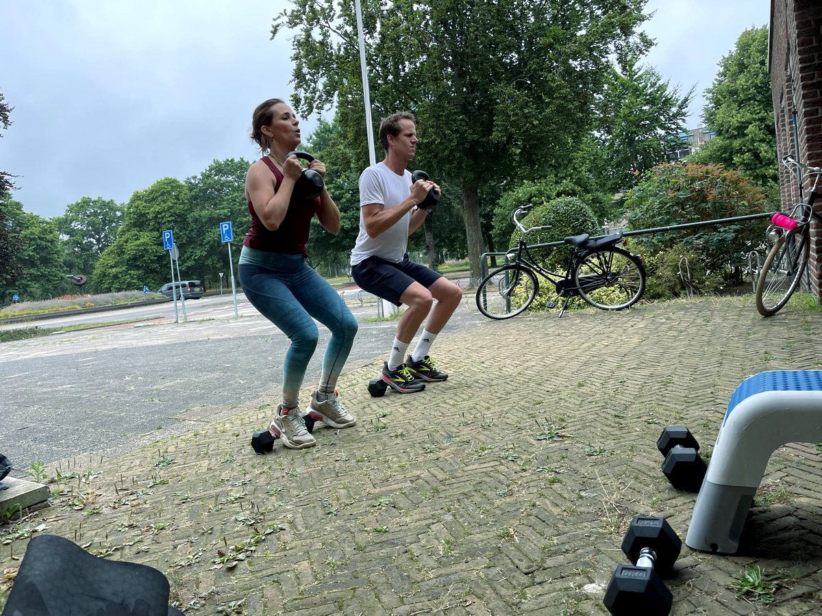 Een man en een vrouw doen tegelijk een kniebuiging met een gewicht op een pleintje